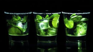 Three glasses filled with lime wedges, mint leaves, ice cubes, and a salted rim, glowing under a dark background