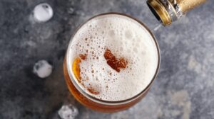 Close-up of light beer being poured into a glass, showing frothy foam and golden color with ice cubes in the background