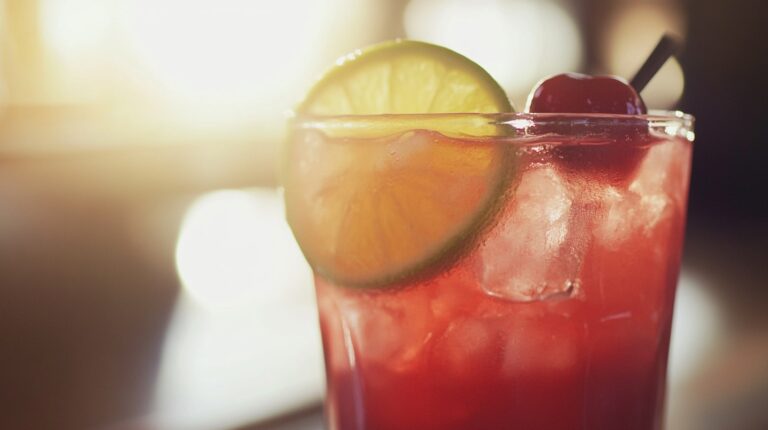 A vibrant red cocktail with ice, garnished with a lime slice and a cherry, glowing in warm sunlight