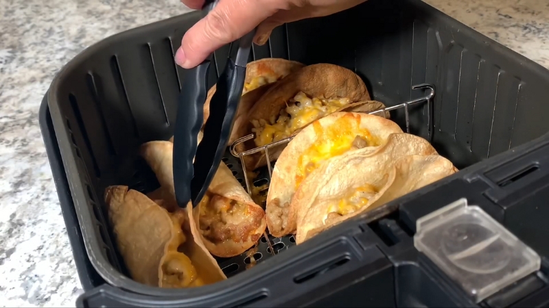 A Hand Using Tongs to Remove Freshly Cooked Taco Shells Filled with Melted Cheese from An Air Fryer Basket