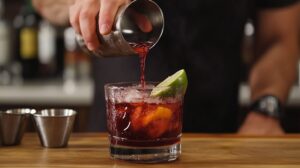 A bartender pours a cherry-colored cocktail from a shaker into a glass garnished with lime and orange slices