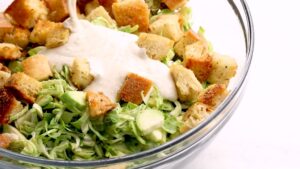 A Glass Bowl Filled with Shredded Brussels Sprouts and Croutons Being Topped with Creamy Caesar Dressing