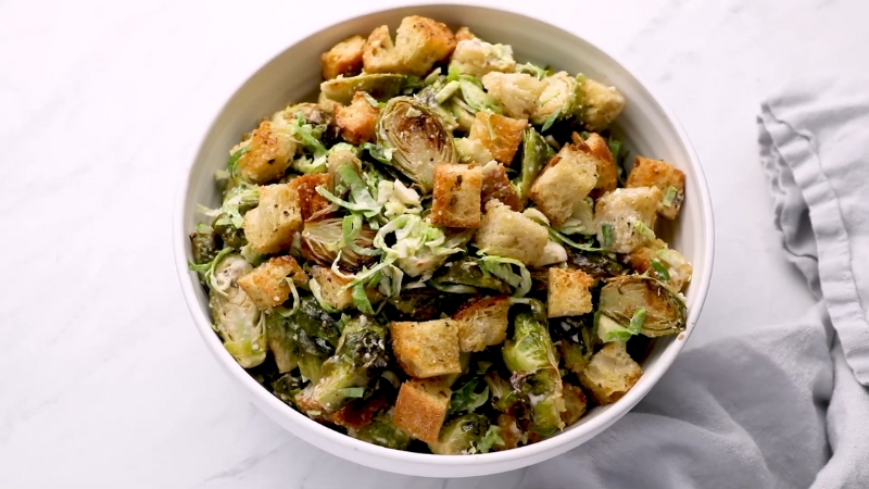 A White Bowl Filled with Crispy Brussels Sprout Caesar Salad Topped with Golden Croutons and Shaved Sprouts on A Marble Surface