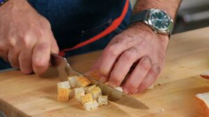 A Person Slicing Fresh Bread Into Small Cubes on A Wooden Cutting Board to Prepare Croutons