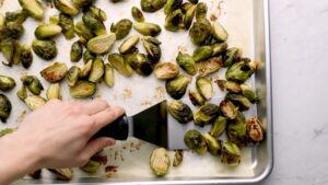 A Hand Using a Spatula to Move Roasted Brussels Sprouts on A Baking Sheet