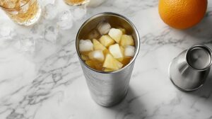 A cocktail shaker filled with ice, pineapple chunks, and juice on a marble countertop with cocktail tools nearby