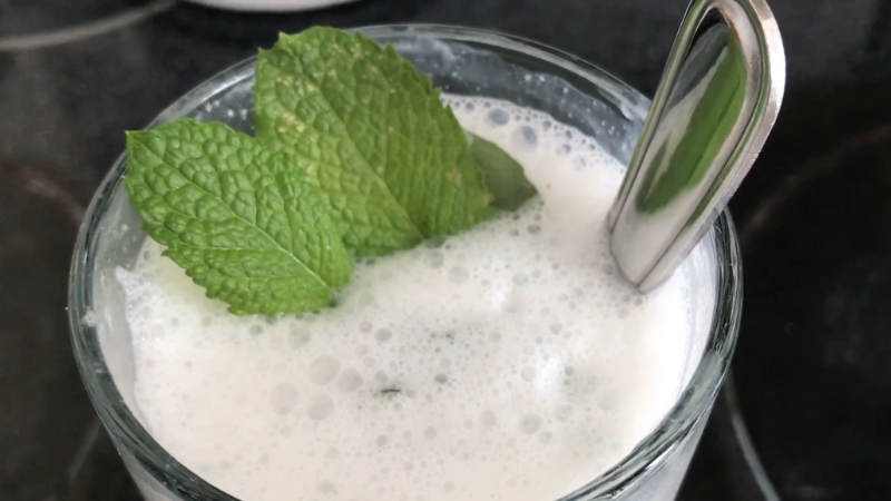 A frothy buttermilk smoothie in a glass, topped with fresh mint leaves and a spoon