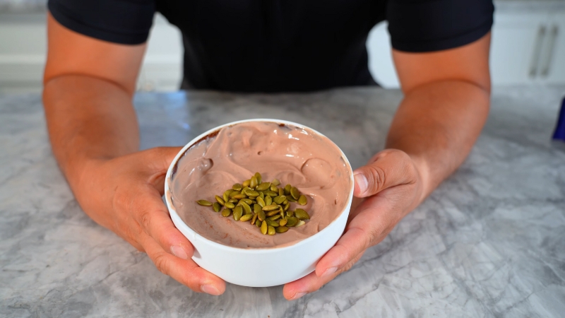 A person holds a bowl of chocolate protein pudding topped with pumpkin seeds