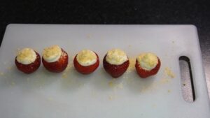 Six filled strawberries topped with crushed graham crackers are arranged on a white cutting board