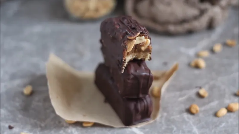 Two stacked no-bake vegan Snickers bars with a bite taken from the top one, showing peanuts and caramel
