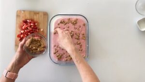 A person sprinkles crushed graham crackers over a pink strawberry mixture in a glass container