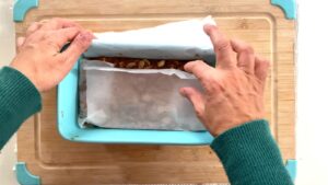 Hands folding parchment paper over peanut caramel mixture in a loaf tin