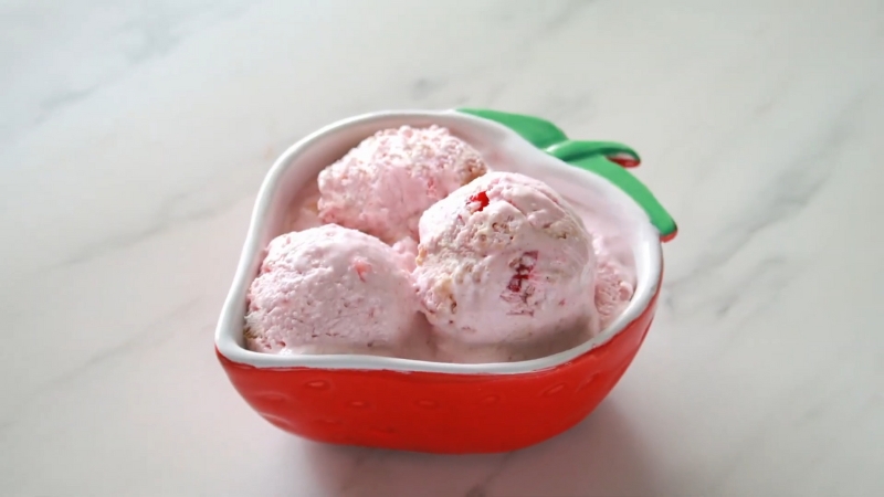 Three scoops of strawberry cheesecake cottage cheese ice cream served in a red, strawberry-shaped bowl on a white marble surface