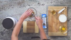 A person mixes ingredients in a bowl surrounded by protein powder, yogurt, honey, and an egg on a countertop