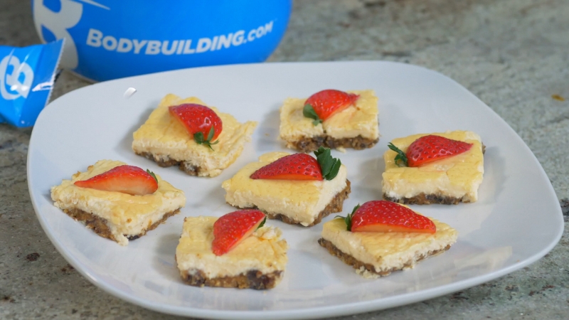 A white plate holds several square no-bake strawberry cheesecake bites topped with halved strawberries