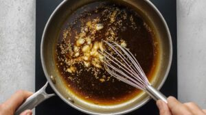Overhead view of a pan with a whisk mixing a garlic butter glaze made of soy sauce, garlic chunks, and sesame seeds