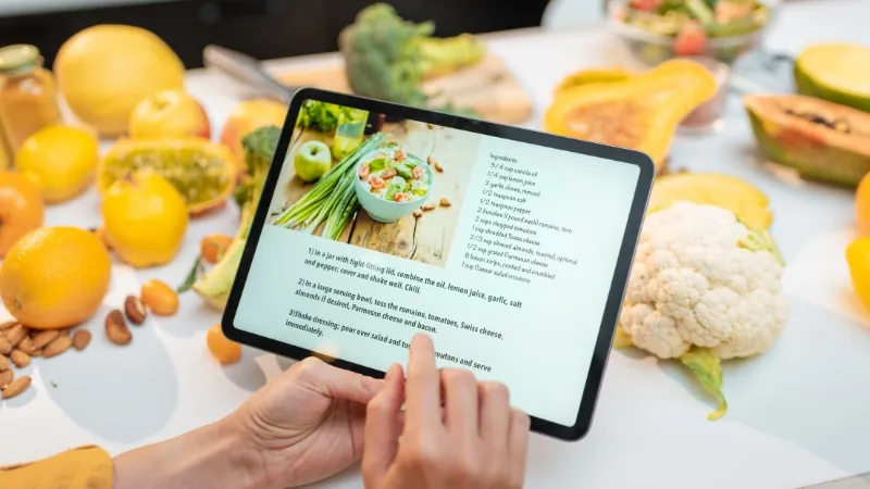 Hands hold a tablet that shows cookbook details beside fresh produce on a table
