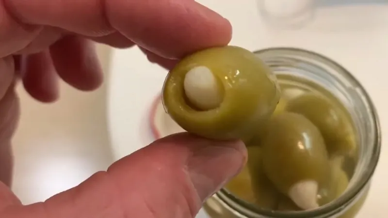 A hand holds a green olive stuffed with garlic above an open jar of garlic-stuffed olives