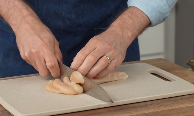 Hands slicing raw chicken breast on a white cutting board