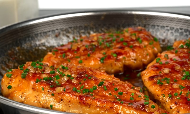 Three grilled chicken pieces with a glossy, brown glaze and chopped green herbs are sizzling in a pan