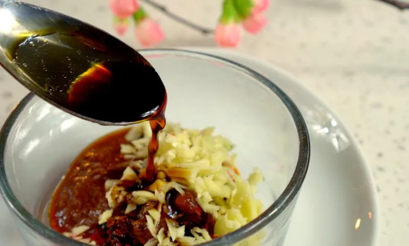 A spoon drizzles dark soy sauce over neatly arranged shredded cheese, chopped nuts, and sauce in a glass bowl