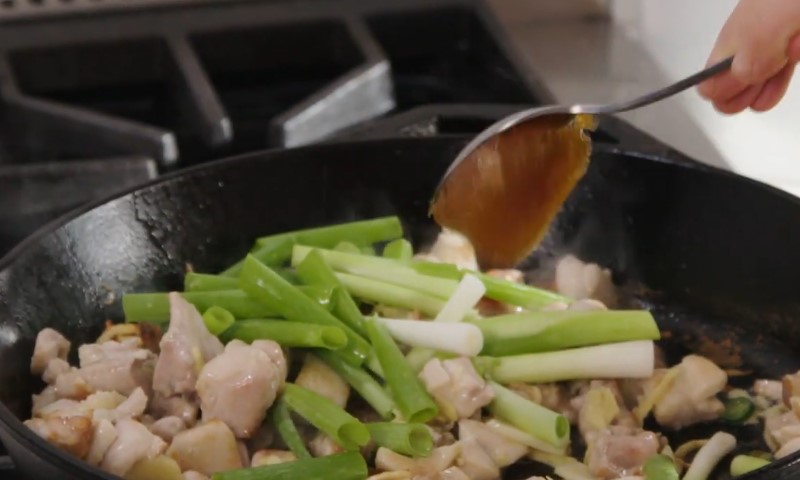 A hand pours sauce from a spoon over cooking chicken pieces and green onions in a black skillet