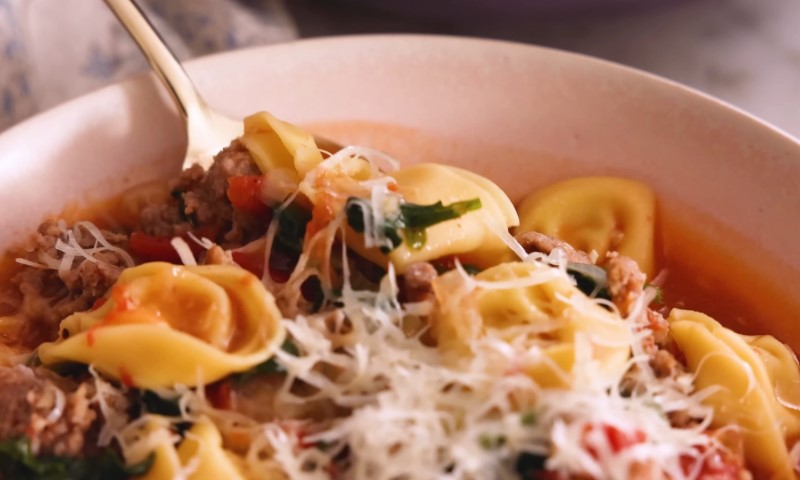 A close-up of a bowl of tortellini soup with ground meat, tomatoes, greens, and grated cheese on top