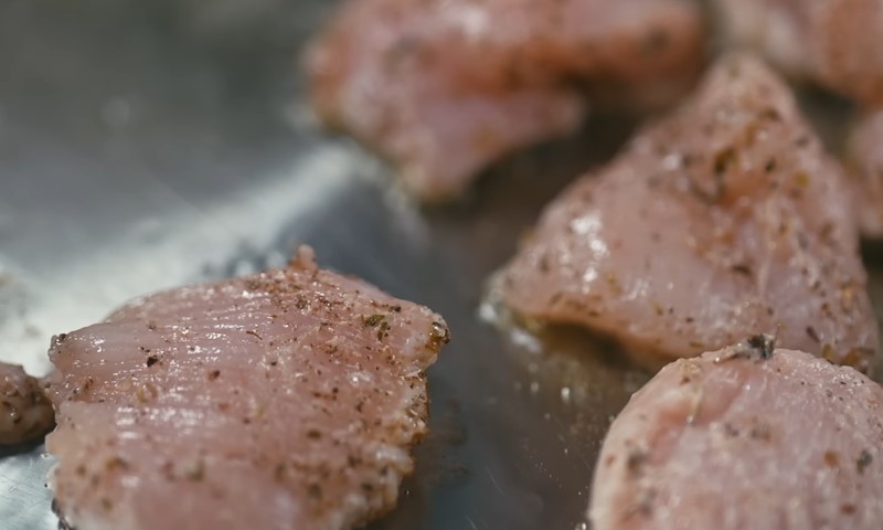 Close-up of raw chicken pieces seasoned with pepper and herbs on a shiny metal surface