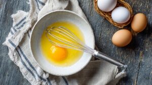 Cracked egg in a bowl with a whisk, surrounded by whole eggs on a rustic wooden surface