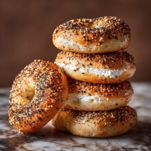 Stack of golden everything bagels, some sliced and spread with cream cheese, on a marble surface
