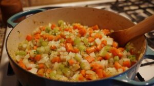 Diced carrots, celery, potatoes, and onions cooking in a pot while being stirred with a wooden spoon