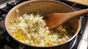 Diced onions cooking in oil with herbs in a pot on a stovetop while being stirred with a wooden spoon