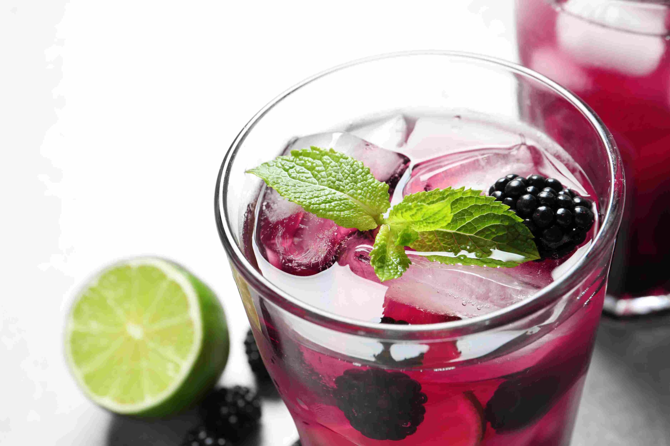 Glass of deep pink berry drink with ice, mint leaves, blackberries, and lime on the side