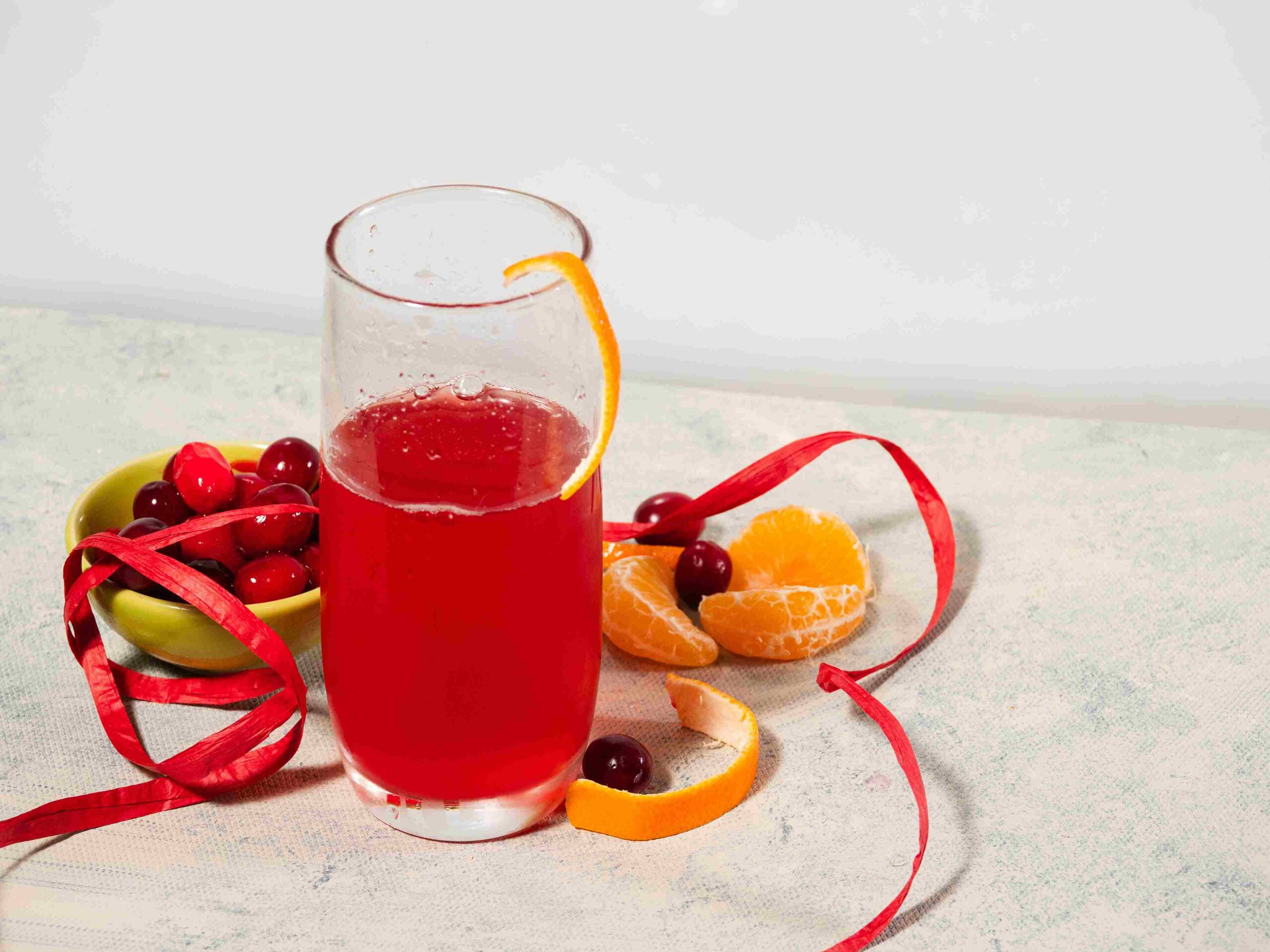 Glass of bright red cranberry drink with orange peel, cranberries, and citrus slices on a table