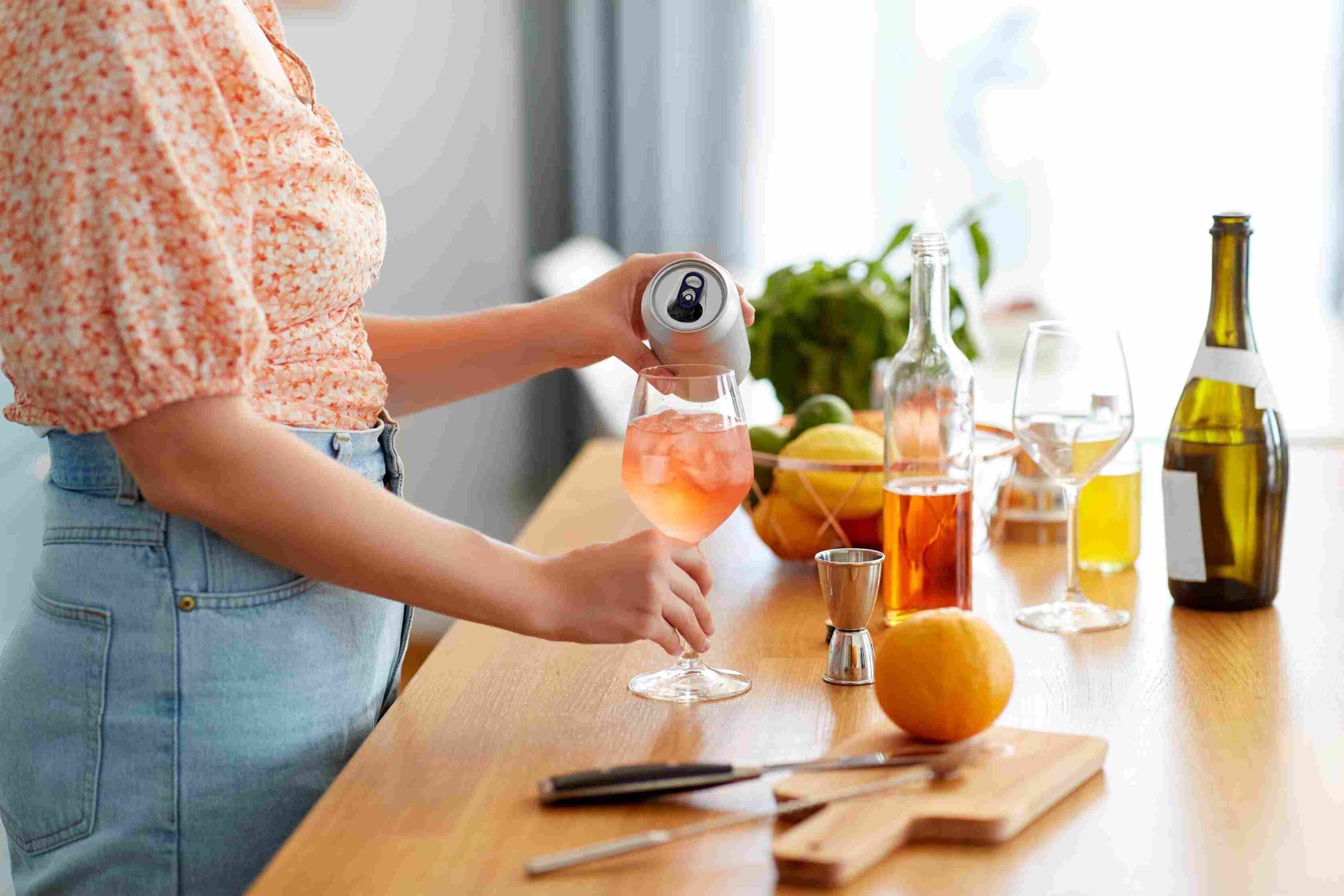 Person pouring a beverage into a glass with bottles and fruit on a kitchen counter