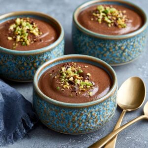 Three bowls of chocolate phirni garnished with chopped pistachios, served on a gray surface with gold spoons