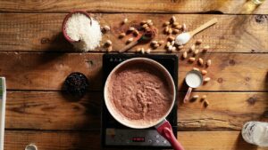 A saucepan of chocolate phirni cooking on a stovetop, surrounded by rice, cocoa powder, almonds, and milk